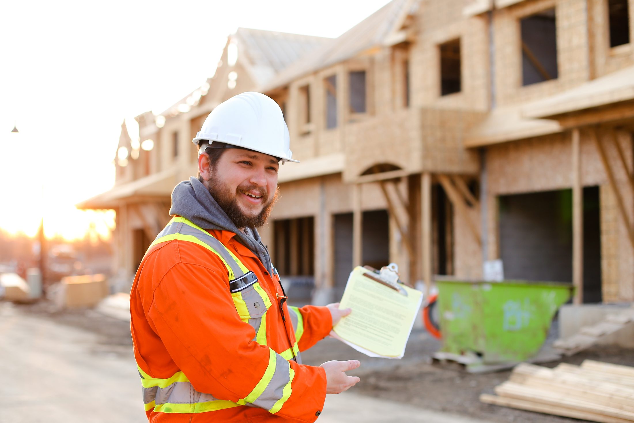A smiling construction professional in safety gear holding a clipboard at a residential building site at sunset