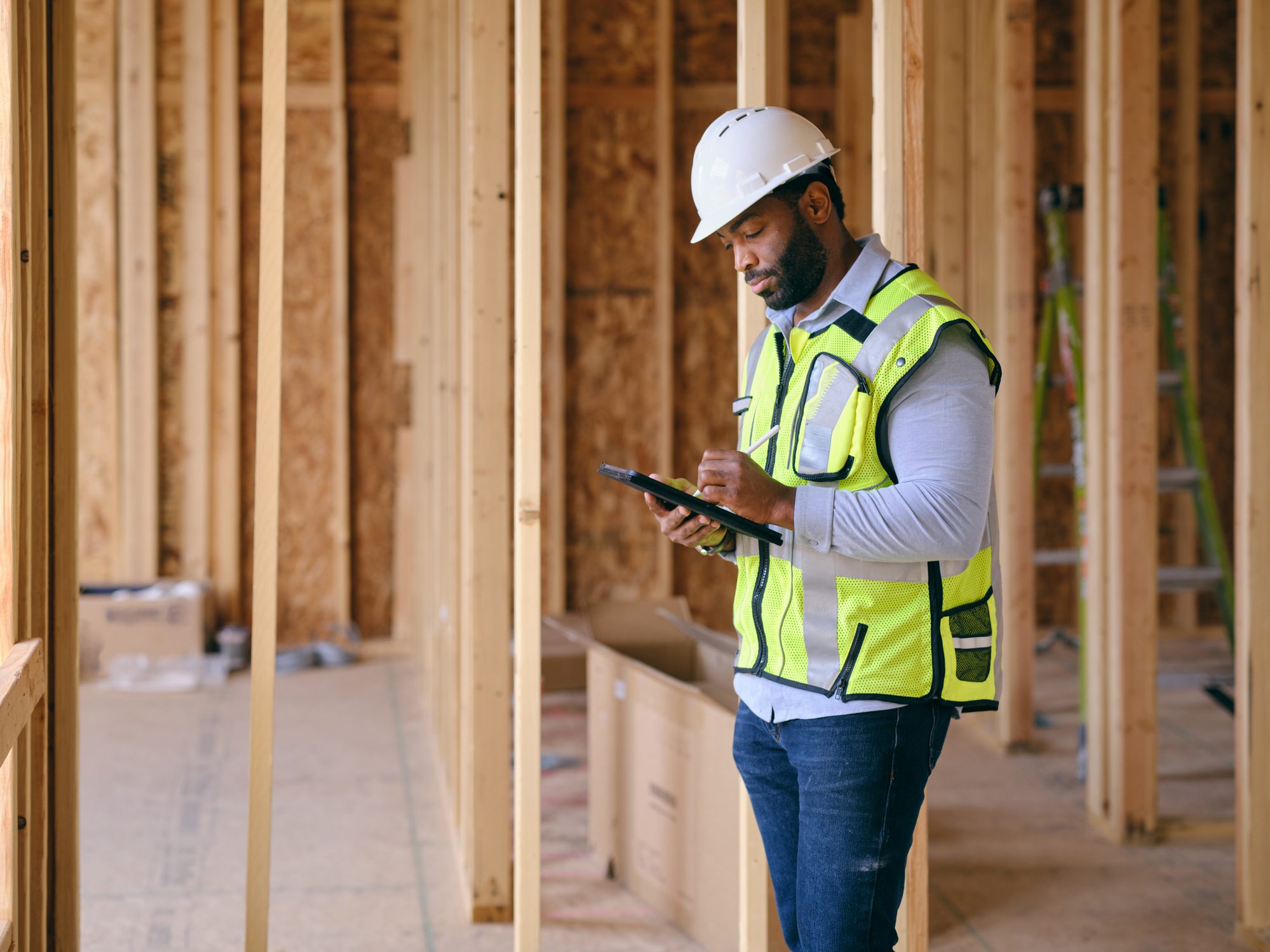 A construction inspector in a high-visibility vest recording notes on a tablet inside a wood-framed building