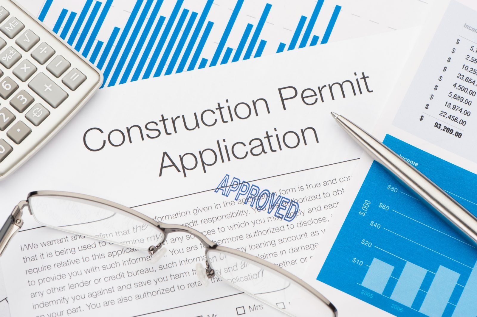 A stamped construction permit application beside a calculator, glasses and summary charts.